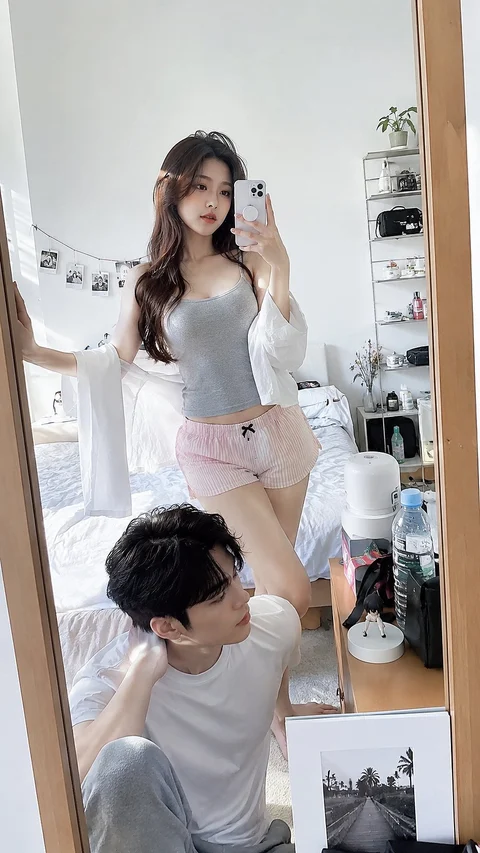 A woman holding a phone stands behind a seated man in a bright room reflected in a tall mirror.