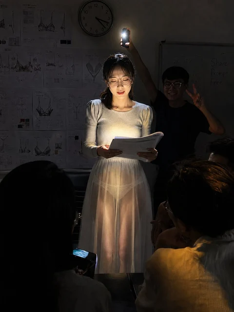 A woman in a pale dress holds an open book illuminated by a small light in a dark room.