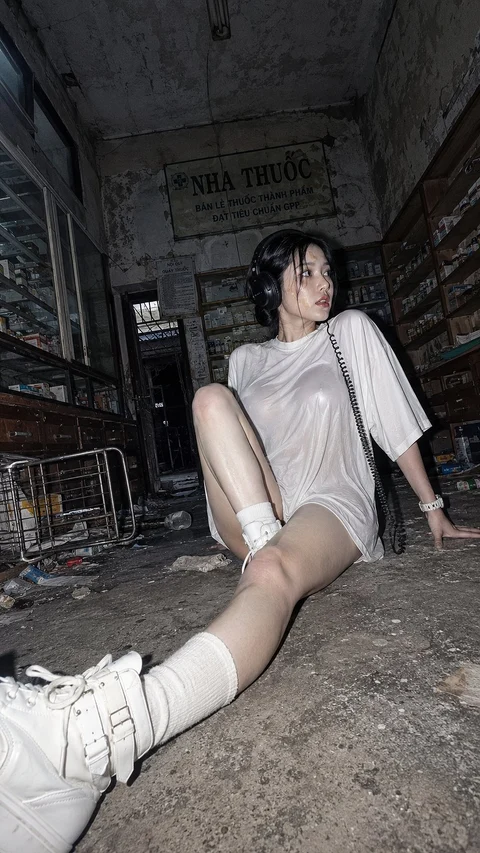 A person in an oversized pale shirt, white socks, and sneakers sits on the floor of a dark, weathered building.